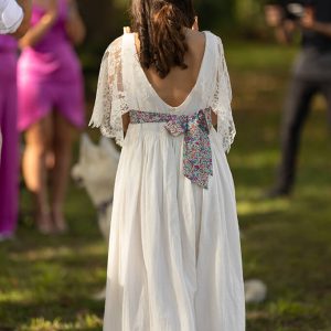 Jeune fille en robe longue blanche pour une cérémonie de mariage style bohème. La robe a le dos en v, des manches en dentelle de Calais et une ceinture en Liberty, elle est en coton brodé et plumetis.