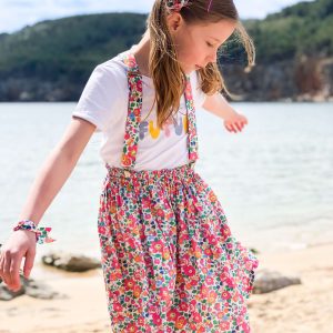 Petite fille portant une jupe et des bretelles en liberty, elle est devant la mer, on voit une colline en arrière plan.