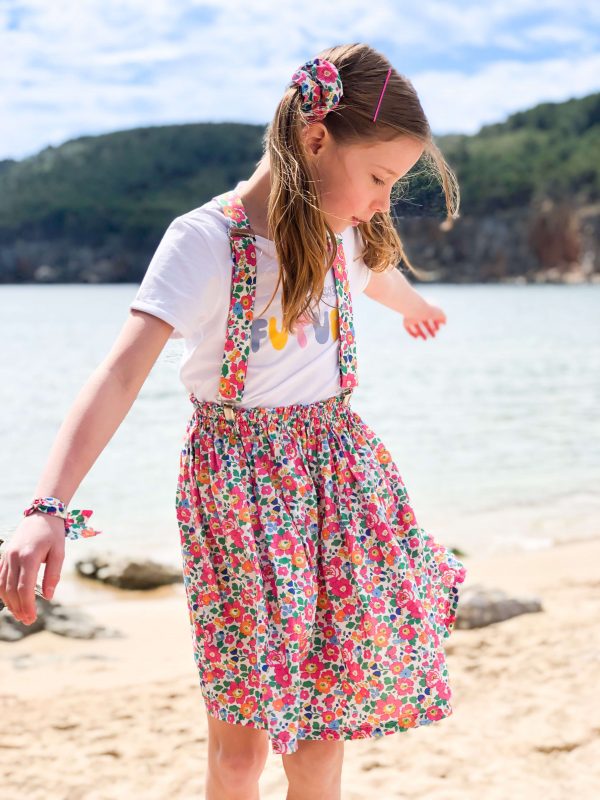 Petite fille portant une jupe et des bretelles en liberty, elle est devant la mer, on voit une colline en arrière plan.