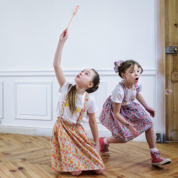 2 petites filles jouant avec des bulles dans un salon. Elles portant des jupes en liberty et des bretelles assorties. Elles ont des converses avec des lacets en liberty, un bracelet en liberty et un chouchou coordonné.