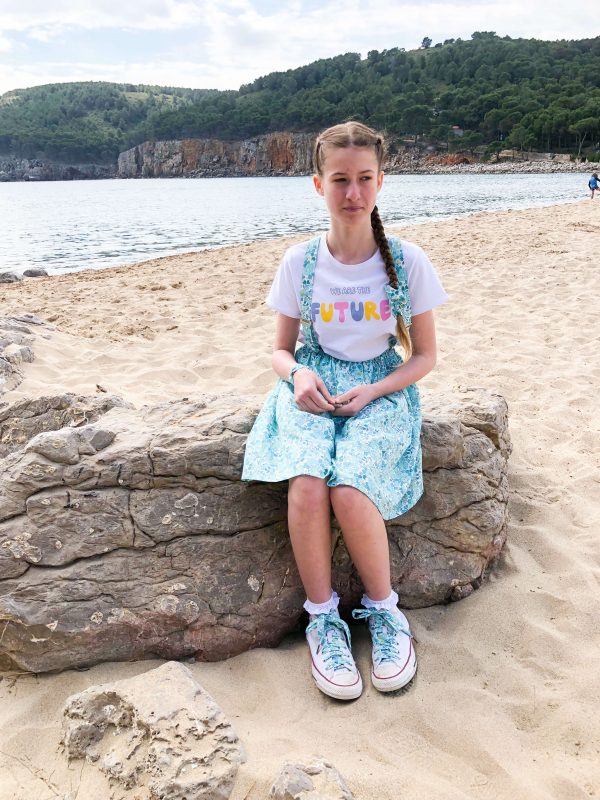 adolescente portant une jupe en liberty, des bretelles et un bracelet assortis. Elle a des converses avec des lacets liberty. Elle est assise sur un rocher sur la plage, on voit une colline en arrière plan.