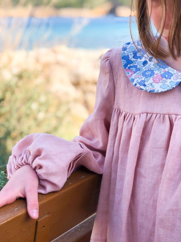 Détail de la manche et du devant de la robe en lin rose à col claudine en liberty, portée par une petite fille. On voit la mer en fond.