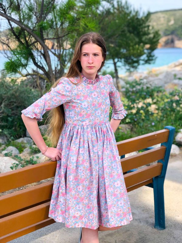 jeune fille romantique portant une robe à col et manches volantés en Liberty Meadow Song pétales. Elle est adossée au banc devant la plage