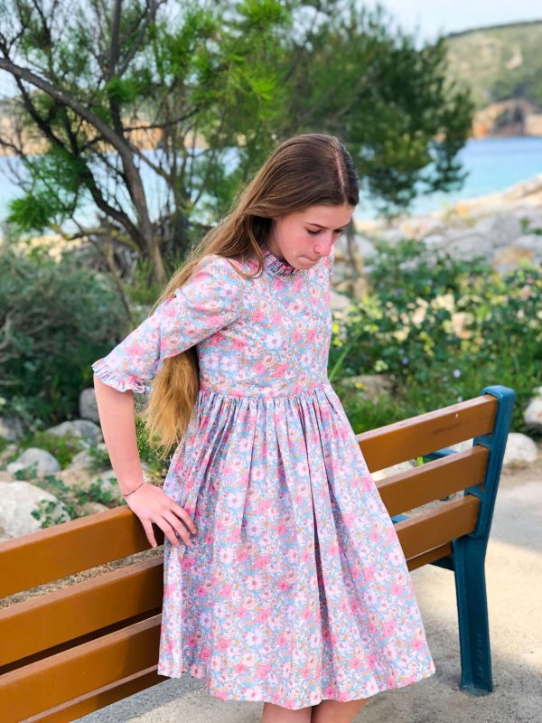 Jeune fille adossée à un banc portant une robe manches longues volantées en liberty meadow song pétales pour fille