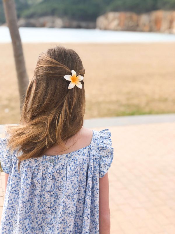 Petite fille blonde aux cheveux longs avec une barrette en forme de fleur dans les cheveux. Elle porte une robe à encollure carrée et manches courtes volantées en liberty bleu. Elle est devant la plage.