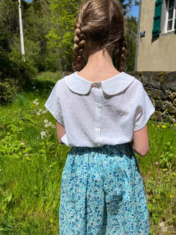 Petite fille portant une jupe en liberty wiltshire blue crystal et une blouse blanche à col claudine en plumetis de coton. Elle a des tresses et les cheveux blonds.