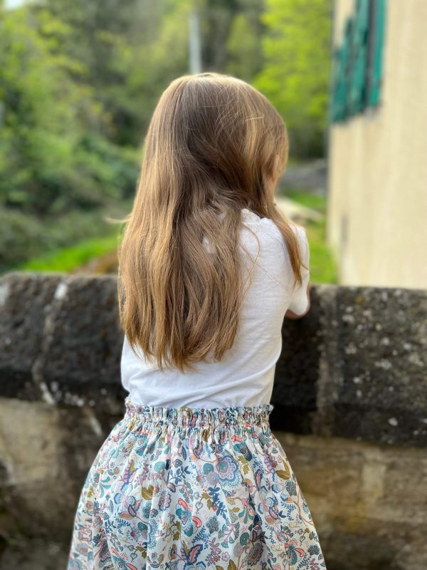 petite fille sur le perron ancien d'une maison, cheveux détaché. Elle porte une jupe en liberty michelle sunflowers et un t-shirt blanc.