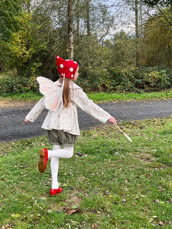 Petite fille portant un ensemble blouse à large col arrondi et bloomer en flanelle kaki, ailes de fée et bonnet de lutin rouge champignon, chaussures rouge lucine